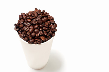 white cardboard Cup with coffee beans on a white empty background. the glass is located on the left side of the frame, the coffee beans are poured into the glass above the edges
