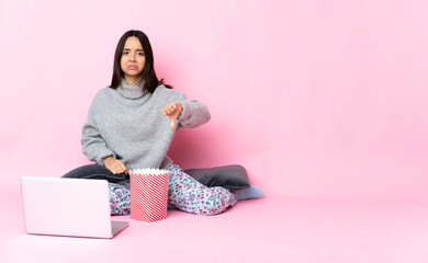 Young mixed race woman eating popcorn while watching a movie on the laptop showing thumb down with negative expression