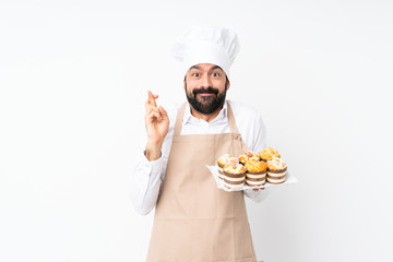 Young man holding muffin cake over isolated white background with fingers crossing and wishing the best