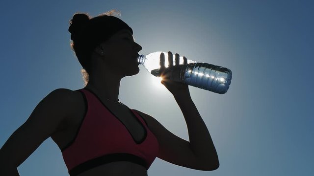Sportwoman drinks water from bottle against sunlight
