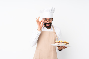 Young man holding muffin cake over isolated white background with glasses and surprised