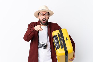 Traveler man man with beard holding a suitcase over isolated white background surprised and pointing front