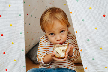 Cute little boy eating bread and butter in a hut, looking up at a camera