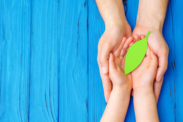 Top view of green paper leaf in hands of adult and child on wooden blue background. Concept of world environment day, world health day and Earth day. Copy space.