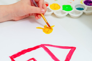 Child's hand drawing yellow sun with red house by watercolors on white sheet of paper. Hand of child drawing the sun.