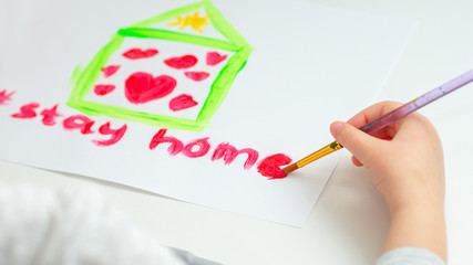 Child is writing words Stay Home with red watercolor under drawn house with hearts on sheet of paper. Stay Home concept.