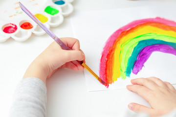 Top view of hands of child drawing a rainbow with brushes and paints on white paper.