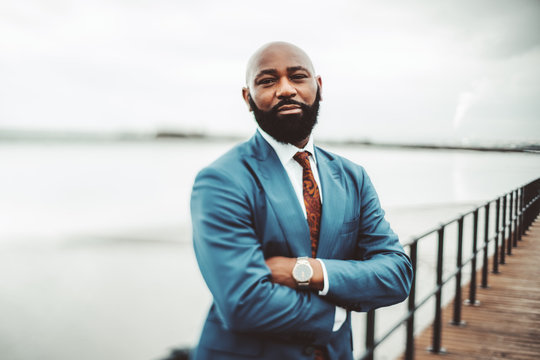 A True Tilt-shift Portrait Of A Successful Adult African Bald Bearded Man In A Blue Suit With Tie And Watch Standing With Hands Crossed On An Embankment Near Facing On An Overcast Day, Selective Focus