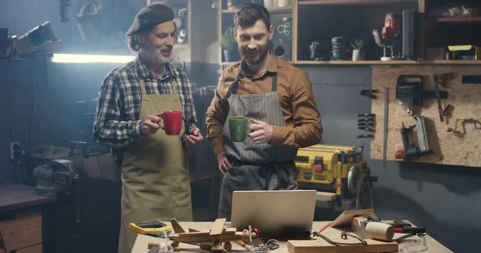 Old and young male carpenters watching video on laptop computer in workshop while smoking and sipping hot drinks. Senior father and son, handicraftsmen resting and drinking coffee or tea in workroom.