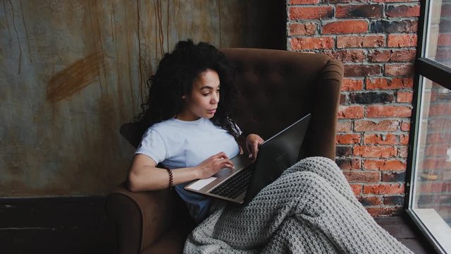 Portrait of young attractive African-American woman using laptop computer sitting in chair in her loft bedroom. Work from home concept