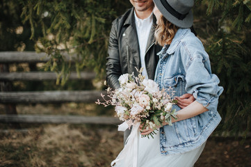 The bride in a denim jacket and hat holds a bouquet in white and purple shades. Groom hugs her from...