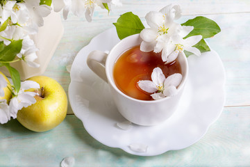 White cup of tea on shaped saucer with blossoming apple tree flowers and yellow apple on wooden table with bokeh closeup. Apple holiday. Apple day. Tea time.