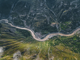 Top down aerial drone view of river and small settlement in between the tall mountains in Tibetan area of Sichuan province, China. Cinematic shot, curved serpentine roads and hills. Journey in spring.