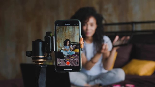 Young african american woman make up blogger streaming live with a smartphone reviewing make up products while sitting on her bed at home, dolly shot