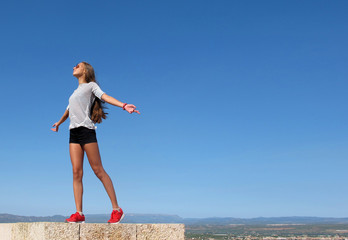 A teenage girl with long hair stands on the stone wall of an old castle with her arms outstretched to meet the wind against a bright blue sky.  Сoncept of freedom and inspiration. Travel content
