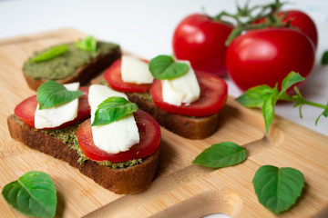 Traditional Italian Caprese bruschetta: sandwich toast with basil pesto sauce, mozzarella cheese and tomatoes on the wooden board surrounded by basil leaves and tomatoes