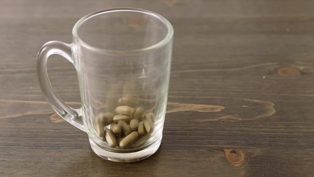 Herbal medicine tablets (black walnut leaves) fall into a transparent empty glass on a wooden table. Concept for self-medication, natural medicine and dangers of overdose
