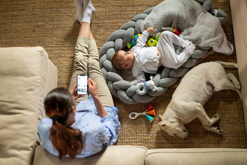 Obraz na płótnie Canvas Authentic top view of an young neo mother is doing smart working with smart phone on a floor with her newborn baby and sleeping puppy in a living room at home.