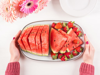 Watermelon slices with strawberries on ceramic tray on white wooden table with pink flowers. Fresh summer snack