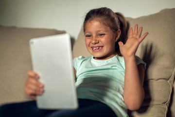 Authentic shot of a happy little girl is having fun to make a selfie or video call with a tablet to parents or relatives while sitting on a sofa in a living room at home.