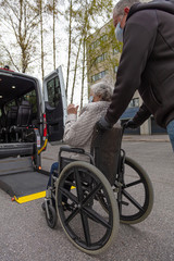 Handicapped person loading in a wheelchair into a minibus for transportation.
Male driver and elderly woman in medical masks.