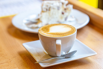 A cup of coffee on a caffe wooden table with piece of cake in the background.