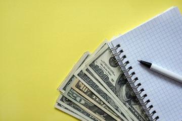 Business elements - money fan, white notebook, ball pen on yellow background. Banknotes of various denominations. Top view. Selective focus. Close-up. Copy space.