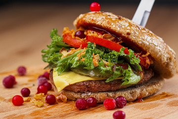 Delicious juicy burger with beef patty, bun, cranberry sauce and vegetables on a serving wooden board on a black background. Food delivery. Fast food restaurant.
