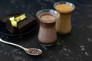 Dalgona coffee and cocoa in glass transparent glasses. Nearby lies a metal spoon with instant coffee, and a chocolate dessert on a saucer