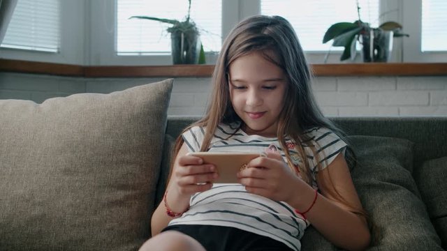 Cut beautiful kid girl plays in a smartphone sitting on the sofa
