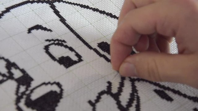 A man embroiders a white thread on a picture. A needle with a thread for embroidering a picture with a cross is clamped in a male hand.
