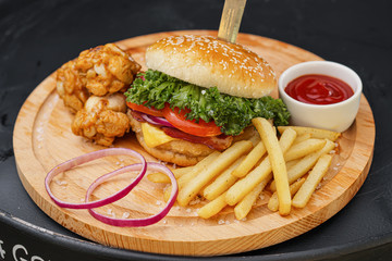 Combo set of fast food. Juicy hamburger, French fries, chicken legs and sauce. Served wooden board and cutlery on a black background. Fast food restaurant. Food delivery.
