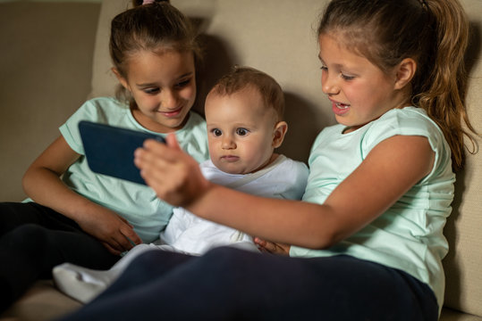 Authentic shot of happy little sisters with newborn brother are making a selfie or video call to parent or relatives while sitting on a sofa in a living room at home. 