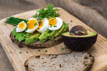 A slice of bread with avocado and eggs on a wooden board. Also salt, chives, parsley, half of avocado and another slice of bread. There is a sackcloth in the background