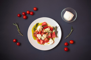 White plate with caprese salad surrounded by cherry tomatoes, mozzarella cheese and rosemary. Top view.