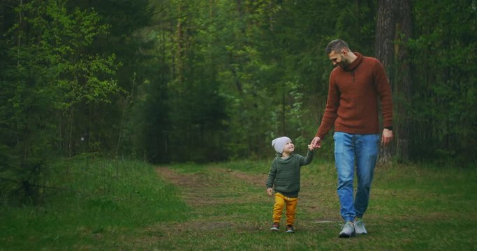 A young father and son walk in the Park holding hands. A man and a boy walk in a pine forest together. Slow motion happy father and son. Fathers day. a 2-year-old boy in the woods with his father.