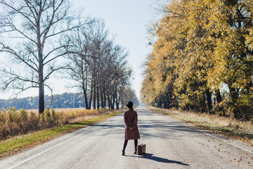 Young redhead lady woman in pink vintage coat and hat with suitcase in retro style walking away along a park road with golden yellow autumnal trees. Outdoor autumn garden relaxation, travel concept