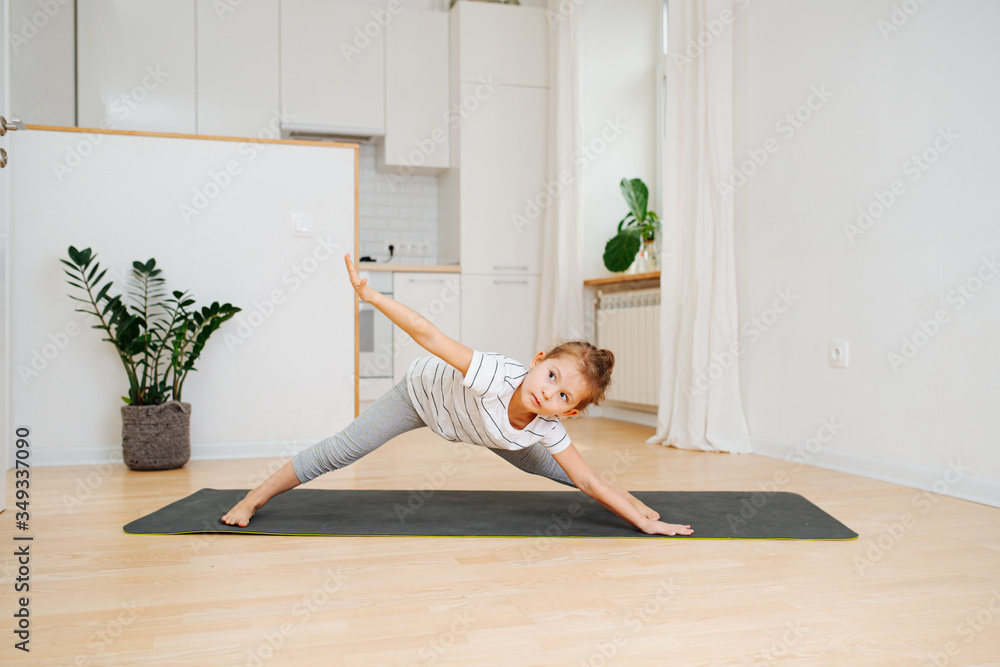 Wall mural Six year old girl doing yoga at home, bending forward with legs wide apart