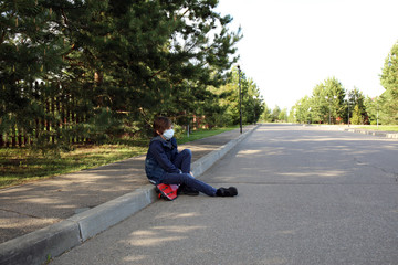A boy walks on the street in a medical mask. A child rides on the road on a red skateboard in a mask.