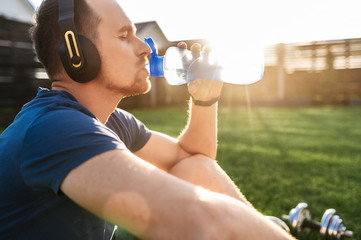 The guy quenches thirst during an outdoor workout. He sits with a headphones and drinks water from...