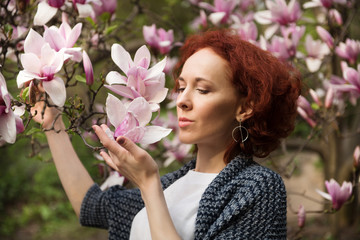 Portrait of young pretty red haired woman near blooming magnolia tree on sunny spring day