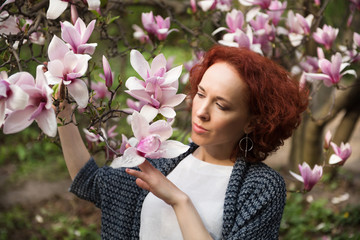 Fototapeta premium Portrait of young pretty red haired woman near blooming magnolia tree on sunny spring day