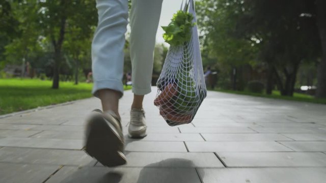 Front view of woman holds cotton mesh shopping bag with vegetables and walks in the summer park