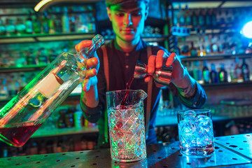 Professional barman finishes preparation of alcoholic cocktail for guest in multicolored neon light. Entertainment, drinks, service concept. Modern bar, crafted beverages, trendy neoned colors.