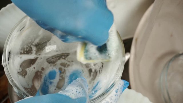 A male hand in a rubber glove washes dishes with a foamy sponge.