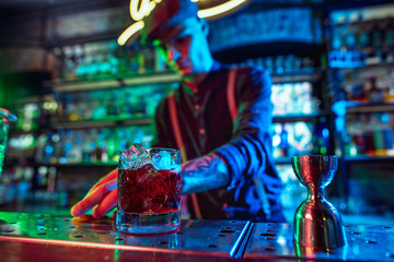 Close up of professional barman finishes preparation of alcoholic cocktail in multicolored neon light, gives it to client. Entertainment, drinks, service concept. Modern bar, trendy neoned colors.