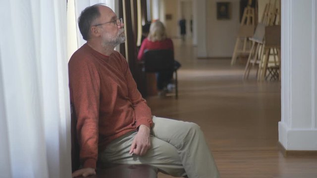 The man in glasses sits on a bench in the gallery and looks with interest at pictures in the hall.