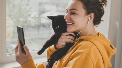 Cheerful young woman wearing headphones holds black pet cat using smartphone for video call,...