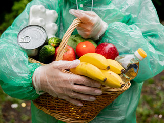 Volunteer food delivery in any weather concept. Portrait of a basket of food in the hands of a woman volunteer in protective gloves and rain coat, close-up.