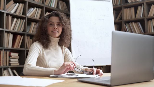 Confident smiling latin hispanic female young school math teacher, college tutor looking at camera sitting at classroom library desk with laptop. School teaching, online education concept. Portrait.